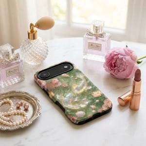 Green koi fish phone case on a white vanity beside perfume bottles, lipstick, a jewelry dish and a pink peony.