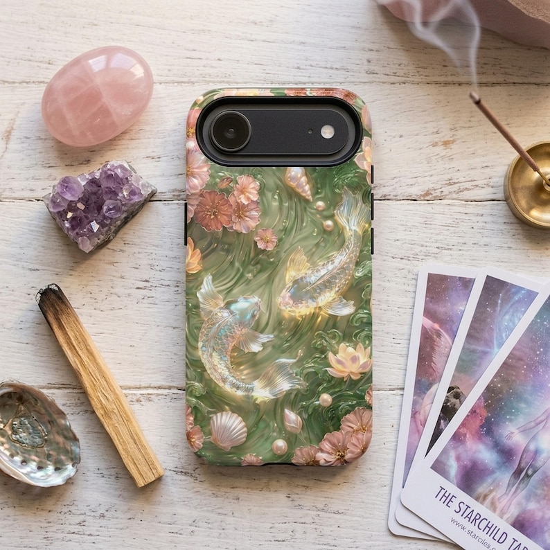 Green koi fish phone case with pink flowers on a white wood table beside rose quartz, amethyst, a wood incense stick and tarot cards.