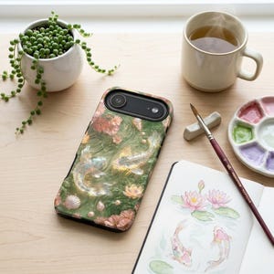 Green koi fish phone case on a white desk beside a potted plant, ceramic mug, watercolor palette, paintbrush and floral sketchbook.