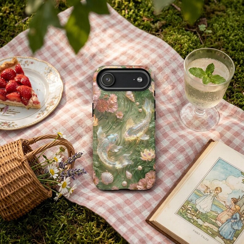 Green koi fish phone case on a pink gingham picnic cloth beside strawberry pie, a mint drink, flowers, a wicker basket and a book.