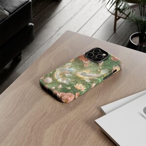 Green floral koi fish phone case on a light wood desk beside the edge of a laptop and a stack of white papers.
