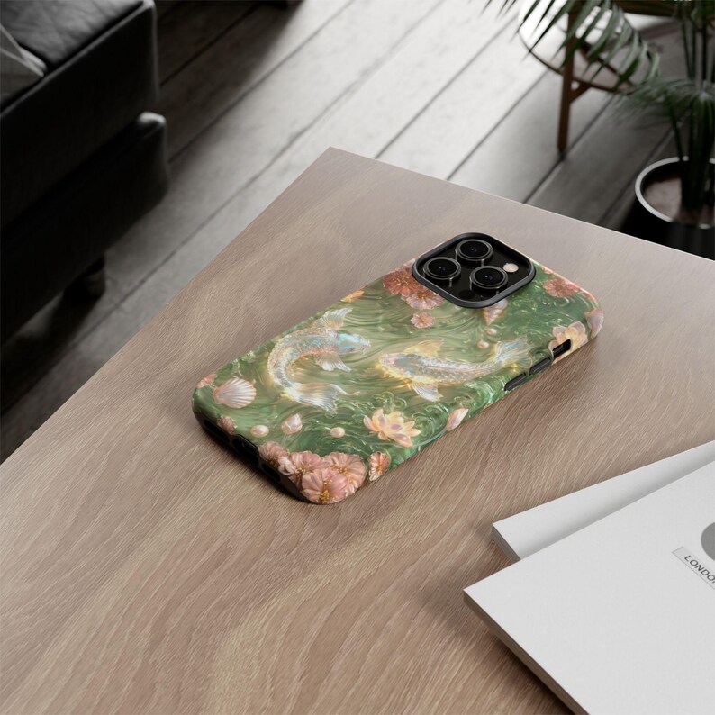 Green floral koi fish phone case on a light wood desk beside the edge of a laptop and a stack of white papers.