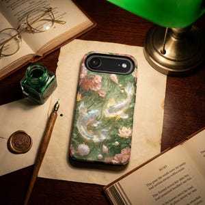 Green floral koi fish phone case on a dark wood desk beneath a banker lamp, beside papers, eyeglasses, a compass and a green stone.