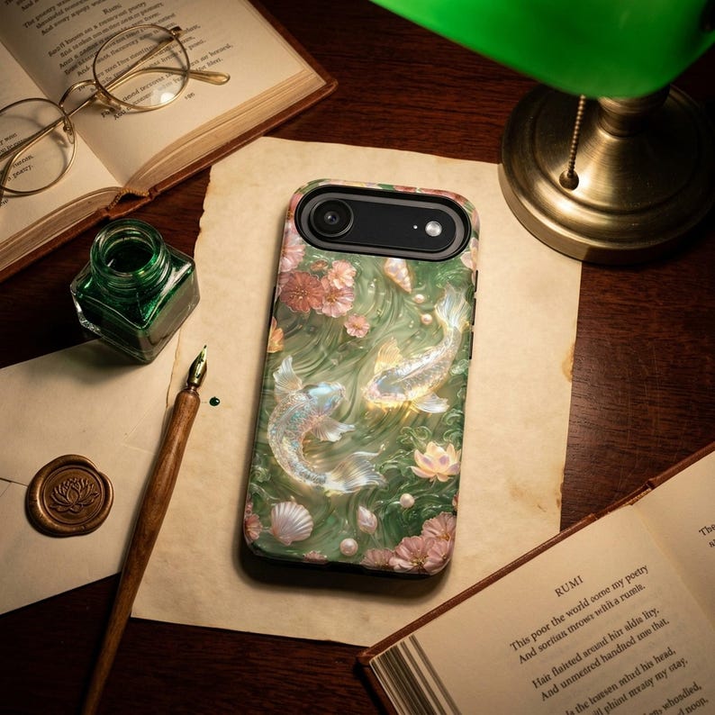 Green floral koi fish phone case on a dark wood desk beneath a banker lamp, beside papers, eyeglasses, a compass and a green stone.