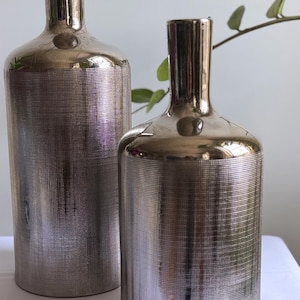 May include: Two cylindrical vases with a metallic silver finish and a fine cross-hatched texture. The taller vase is positioned behind the shorter, wider vase. Both feature a smooth, reflective neck and a flat base, creating a uniform appearance.