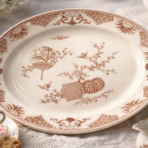 May include: Oval serving platter with a cream-colored base and a brown floral and oriental design. The platter features a decorative border and a central illustration of a fan, birds, and flowering branches. A classic piece for serving.