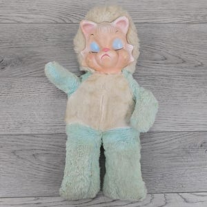 May include: Vintage plush doll with a lion-like face and a light blue and cream-colored body. The doll has a plastic face with closed blue eyes and a sad expression. The plush fur is worn, giving it a nostalgic feel.
