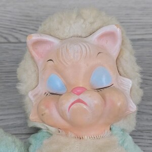 Op de afbeelding: Een vintage poppenhoofd met een leeuwachtig gezicht. De pop heeft gesloten blauwe ogen, een roze neus en een licht perzikkleurige teint. De manen van de pop zijn gemaakt van een zacht, gebroken wit materiaal en de pop draagt een lichtblauwe en cr&egrave;mekleurige outfit.