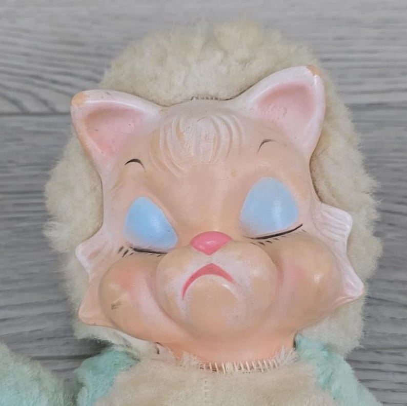 Op de afbeelding: Een vintage poppenhoofd met een leeuwachtig gezicht. De pop heeft gesloten blauwe ogen, een roze neus en een licht perzikkleurige teint. De manen van de pop zijn gemaakt van een zacht, gebroken wit materiaal en de pop draagt een lichtblauwe en cr&egrave;mekleurige outfit.