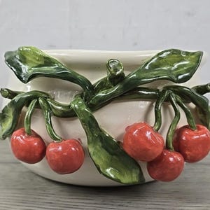 May include: Cream-colored ceramic bowl with a cherry branch design. The bowl features bright red cherries and green leaves. The cherries and leaves are sculpted and attached to the outside of the bowl, creating a three-dimensional effect.