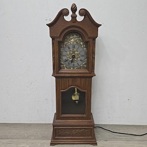 May include: A wooden grandfather clock with a decorative top and a pendulum visible through a glass door. The clock face has Roman numerals and ornate detailing. The clock is brown and has a power cord.