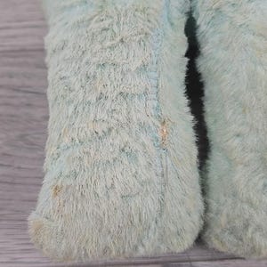 Op de afbeelding: Close-up van het onderste deel van een lichtblauw pluche speelgoed. De vacht van het speelgoed is lang en harig, met zichtbare stiksels en lichte verkleuring. De afbeelding richt zich op de textuur en kleur van de stof.