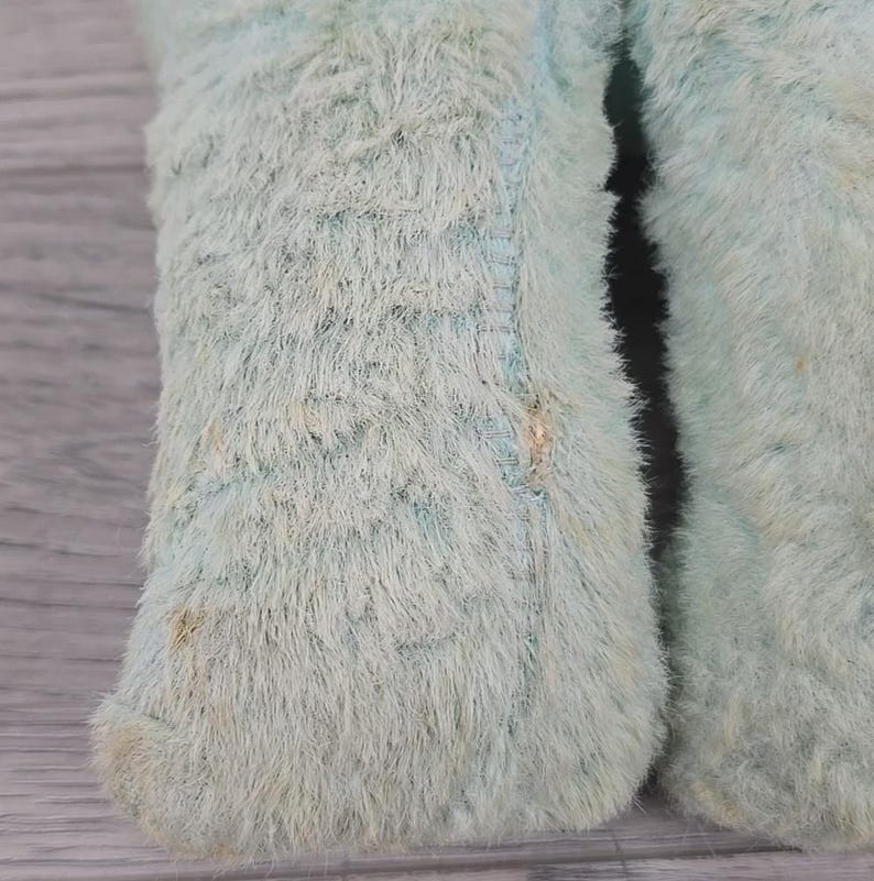 Op de afbeelding: Close-up van het onderste deel van een lichtblauw pluche speelgoed. De vacht van het speelgoed is lang en harig, met zichtbare stiksels en lichte verkleuring. De afbeelding richt zich op de textuur en kleur van de stof.