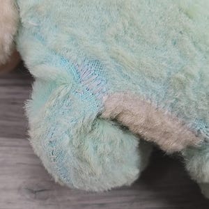 Op de afbeelding: Close-up van de poot en het lichaam van een pluchen speelgoed. Het speelgoed is een mix van pastelkleuren, waaronder lichtblauw, roze en wit. Zichtbare stiksels duiden op een reparatie. De vacht is zacht en pluizig en vertoont tekenen van slijtage.