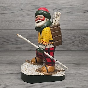 May include: A carved wooden figurine of a skier, wearing a red hat, green scarf, and yellow jacket. The skier has a white beard and is carrying a backpack with birch logs. The skier is wearing red and black plaid pants and brown boots with snowshoes.