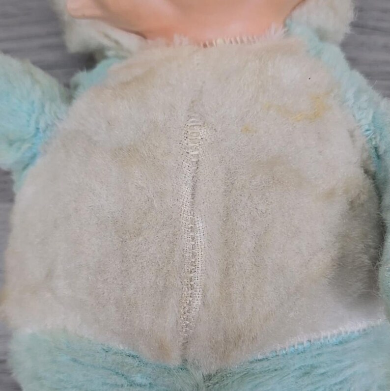Op de afbeelding: Close-up van een vintage pluche speelgoed. Het speelgoed heeft een lichtblauwe buitenkant met een wit, pluizig midden. Het midden heeft een zichtbare naad in het midden. Er zitten wat gele vlekken op het witte bont. Het gezicht van het speelgoed is bovenaan zichtbaar.