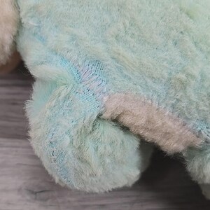 Op de afbeelding: Close-up van de poot en het lichaam van een pluchen speelgoed. Het speelgoed is een mix van pastelkleuren, waaronder lichtblauw, roze en wit. Zichtbare stiksels duiden op een reparatie. De vacht is zacht en pluizig en vertoont tekenen van slijtage.