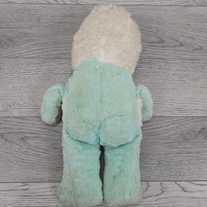 Op de afbeelding: Een vintage pluche speelgoed, gezien vanaf de achterkant, met een lichtblauw lichaam en een cr&egrave;mekleurig hoofd. Het speelgoed heeft een zachte, fuzzy textuur en een eenvoudig ontwerp, met zichtbare stiksels langs de naden. Het is waarschijnlijk een verzamelobject of een nostalgisch item.