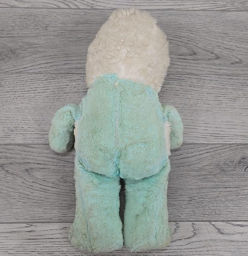 Op de afbeelding: Een vintage pluche speelgoed, gezien vanaf de achterkant, met een lichtblauw lichaam en een cr&egrave;mekleurig hoofd. Het speelgoed heeft een zachte, fuzzy textuur en een eenvoudig ontwerp, met zichtbare stiksels langs de naden. Het is waarschijnlijk een verzamelobject of een nostalgisch item.