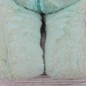 Op de afbeelding: Close-up van het onderlichaam van een vintage, lichtblauw pluche speelgoed. De poten van het speelgoed zijn zichtbaar en tonen een zachte, fuzzy textuur met wat slijtage en verkleuring. Het speelgoed rust op een lichtgekleurd oppervlak.