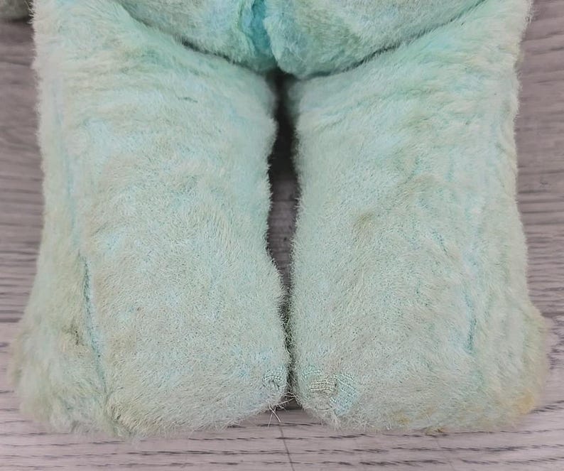 Op de afbeelding: Close-up van het onderlichaam van een vintage, lichtblauw pluche speelgoed. De poten van het speelgoed zijn zichtbaar en tonen een zachte, fuzzy textuur met wat slijtage en verkleuring. Het speelgoed rust op een lichtgekleurd oppervlak.