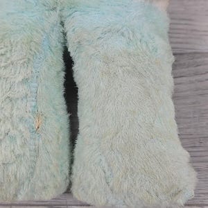 Op de afbeelding: Close-up van een lichtblauw pluche speelgoed met een harige textuur. Het speelgoed vertoont tekenen van slijtage, met zichtbare schade, waaronder een kleine scheur. De stof is vervaagd, wat duidt op leeftijd of gebruik. Het ontwerp van het speelgoed is niet volledig zichtbaar.