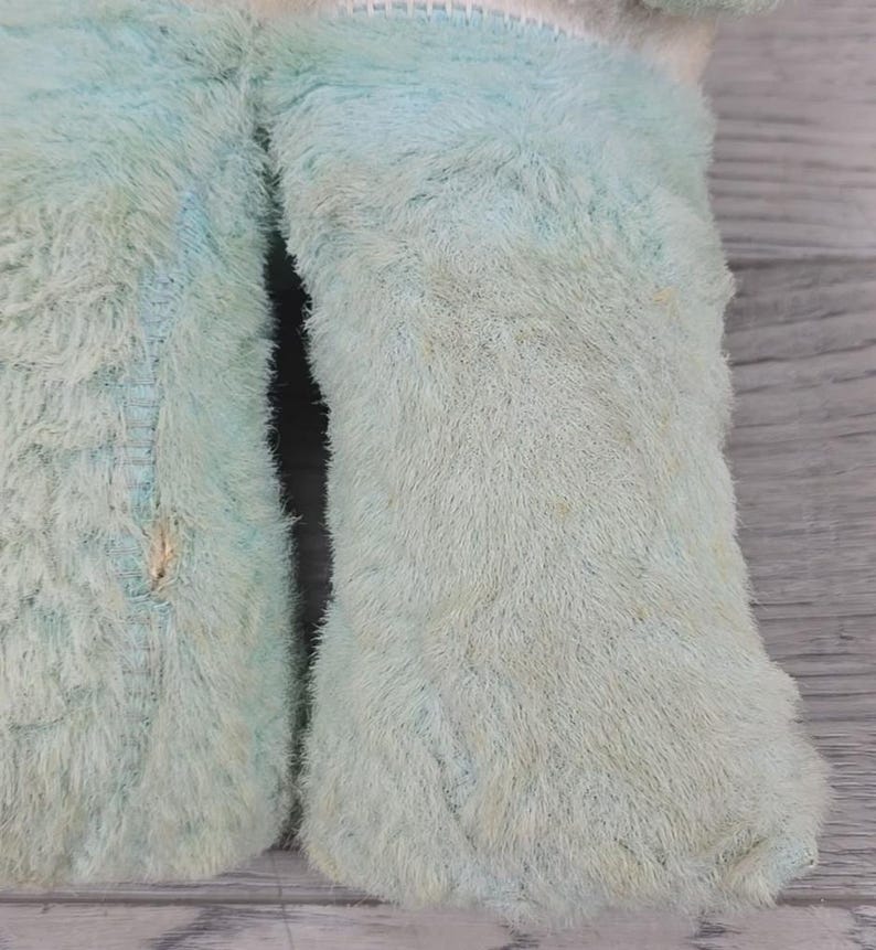 Op de afbeelding: Close-up van een lichtblauw pluche speelgoed met een harige textuur. Het speelgoed vertoont tekenen van slijtage, met zichtbare schade, waaronder een kleine scheur. De stof is vervaagd, wat duidt op leeftijd of gebruik. Het ontwerp van het speelgoed is niet volledig zichtbaar.