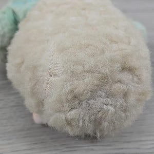 Op de afbeelding: Close-up van een vintage knuffel, met een zacht, cr&egrave;mekleurig lichaam met een getextureerd, pluche oppervlak. Het speelgoed heeft een lichtgroen accent en een klein roze detail. De stiksels zijn zichtbaar aan de zijkant, wat wijst op de handgemaakte aard.
