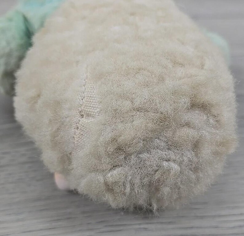 Op de afbeelding: Close-up van een vintage knuffel, met een zacht, cr&egrave;mekleurig lichaam met een getextureerd, pluche oppervlak. Het speelgoed heeft een lichtgroen accent en een klein roze detail. De stiksels zijn zichtbaar aan de zijkant, wat wijst op de handgemaakte aard.