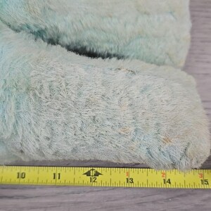 Op de afbeelding: Close-up van een lichtblauw, pluizig pluche speelgoed. De vacht van het speelgoed is versleten en vertoont tekenen van ouderdom. Een gele meetlint is naast het speelgoed geplaatst, metingen van 25,4 cm tot 38,1 cm.