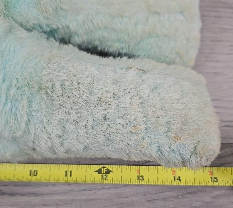 Op de afbeelding: Close-up van een lichtblauw, pluizig pluche speelgoed. De vacht van het speelgoed is versleten en vertoont tekenen van ouderdom. Een gele meetlint is naast het speelgoed geplaatst, metingen van 25,4 cm tot 38,1 cm.