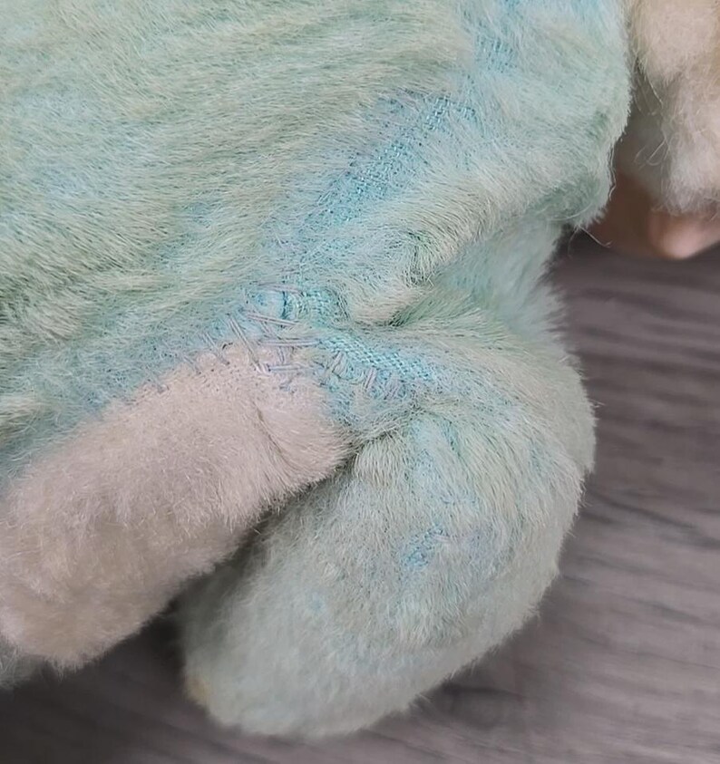Op de afbeelding: Close-up van de poot en het lichaam van een pluche speelgoed. Het speelgoed is gemaakt van zacht, pluizig materiaal in lichtblauw en wit. Zichtbare stiksels duiden op een reparatie. De vacht van het speelgoed heeft een licht versleten uiterlijk.