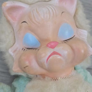 Op de afbeelding: Een vintage kattenkop-pop met gesloten blauwe ogen, een roze neus en mond en roze wangen. De pop heeft een licht perzikkleurig gezicht en een pluizige, gebroken witte manen. Het lichaam van de pop is van zacht, lichtblauw en wit materiaal.