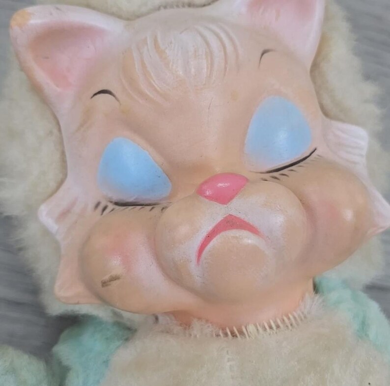 Op de afbeelding: Een vintage kattenkop-pop met gesloten blauwe ogen, een roze neus en mond en roze wangen. De pop heeft een licht perzikkleurig gezicht en een pluizige, gebroken witte manen. Het lichaam van de pop is van zacht, lichtblauw en wit materiaal.