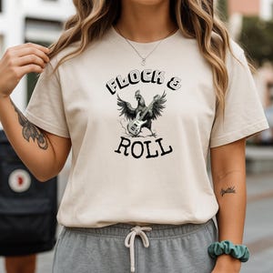 Könnte beinhalten: Beiges T-Shirt mit dem Text "FLOCK & ROLL" über und unter einer schwarz-weißen Illustration eines Gitarre spielenden Huhns. Das Shirt hat kurze Ärmel und einen Rundhalsausschnitt.