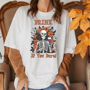 Puede incluir: Camiseta blanca con un diseño de esqueleto sosteniendo una copa de vino, rodeado de flores naranjas. El texto "DRINK IF YOU DARE!" está impreso encima y debajo del diseño del esqueleto. La camiseta tiene mangas cortas y cuello redondo.