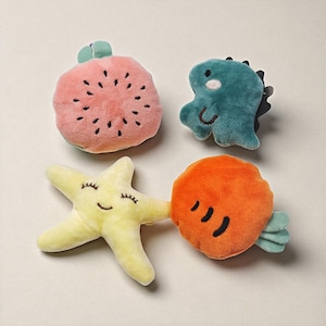 May include: Four plush toys in the shape of a watermelon, dinosaur, starfish, and carrot. The watermelon is pink with black seeds, the dinosaur is teal, the starfish is yellow, and the carrot is orange. Each toy has embroidered details and a soft texture.