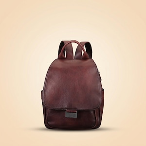 May include: A dark burgundy backpack with a textured surface, featuring a large front flap that covers the main compartment, a rectangular metal clasp closure, and two adjustable shoulder straps attached to the top of the bag.