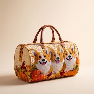 Op de afbeelding: Een gewatteerde reistas met een borduursel van drie Corgi-honden, omringd door oranje pompoenen en herfstbladeren. De tas heeft een crèmekleurige gewatteerde stoffen body, twee bruine lederen handvatten en een ritssluiting aan de bovenkant.