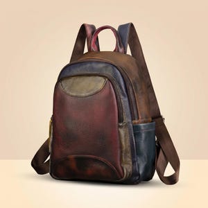 Puede incluir: Una mochila de cuero multicolor con un acabado texturizado en tonos profundos de burdeos, azul marino y verde oliva. La bolsa incluye un asa superior, dos correas ajustables para los hombros, un bolsillo frontal con cremallera y un compartimento.