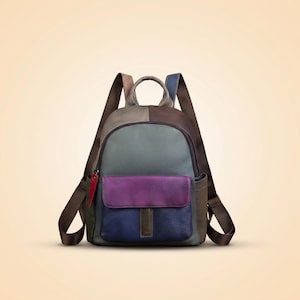 May include: A multi-coloured leather backpack featuring a large grey main compartment, a purple and blue front pocket with a vertical tab, brown adjustable shoulder straps, a top handle, and a small red accent tag on the side zip pull.