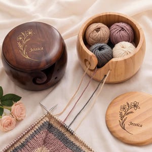 Peut inclure: Deux bols à laine ronds en bois, un brun foncé et un beige clair, chacun doté d'un couvercle gravé d'un motif floral et du nom Jessica. Le bol clair contient quatre petites pelotes de laine beige, mauve, grise et crème, reliées au tricot.