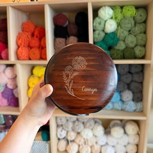 May include: A round, wooden box with a dark brown finish. The lid features an engraved floral design and the name "Camryn". The box is held in front of a wooden shelf filled with colorful yarn skeins, suggesting a craft or storage theme.