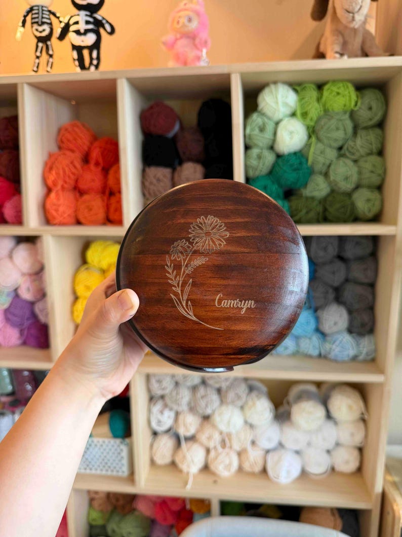 May include: A round, wooden box with a dark brown finish. The lid features an engraved floral design and the name "Camryn". The box is held in front of a wooden shelf filled with colorful yarn skeins, suggesting a craft or storage theme.