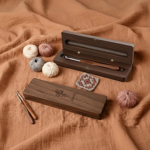 Peut inclure: Un ensemble de crochets en bois avec une gravure de nom personnalisée, présenté dans un étui en bois ouvert. L'ensemble comprend un crochet, un petit carré au crochet et plusieurs pelotes de laine de différentes couleurs. L'étui est gravé d'une rose.
