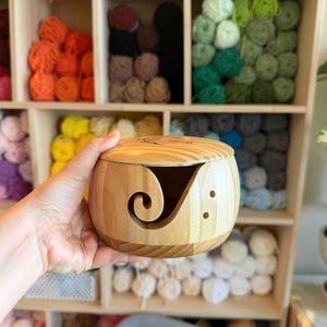May include: A light-colored wooden yarn bowl with a lid. The bowl has a curved slot for yarn and two small holes. The background shows a wooden shelf filled with colorful yarn balls in various shades of orange, green, and blue.