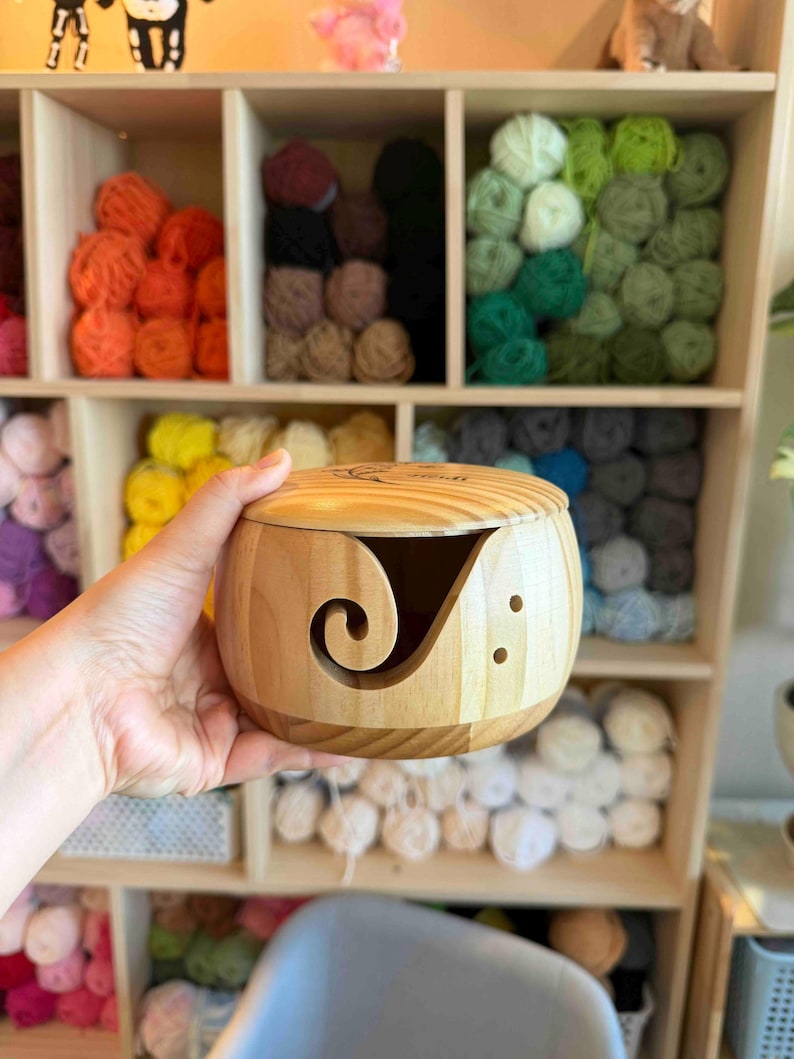 May include: A light-colored wooden yarn bowl with a lid. The bowl has a curved slot for yarn and two small holes. The background shows a wooden shelf filled with colorful yarn balls in various shades of orange, green, and blue.
