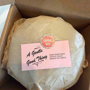 May include: A round, wrapped item in a cardboard box. A pink card with the text "A Gentle Good Thing" and "Hope something gentle today makes you smile" is on top, along with a "Thank You" sticker.