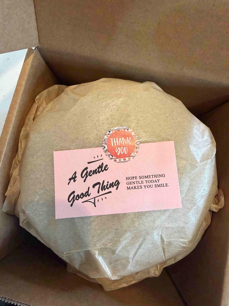 May include: A round, wrapped item in a cardboard box. A pink card with the text "A Gentle Good Thing" and "Hope something gentle today makes you smile" is on top, along with a "Thank You" sticker.