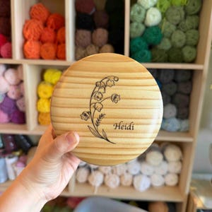 May include: A round, wooden box lid with a floral design and the name "Heidi" engraved on it. The lid is light brown with visible wood grain. The background shows shelves filled with colorful yarn balls.